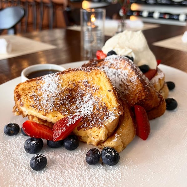 Golden French toast with powdered sugar, strawberries, blueberries, whipped cream, and syrup on a white plate.