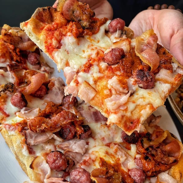 A hand holds up a slice of La Bella Pinseria Romana's meat-topped pinsa, with more pinsa below.