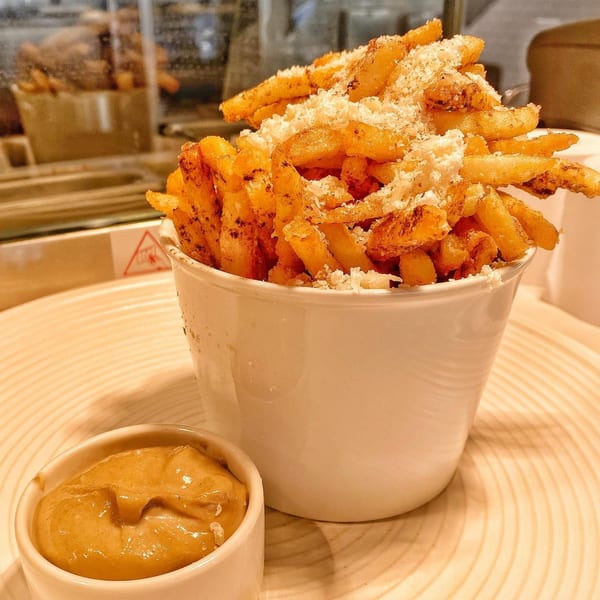A white cup overflowing with golden french fries topped with parmesan cheese, next to a small dish of sauce.