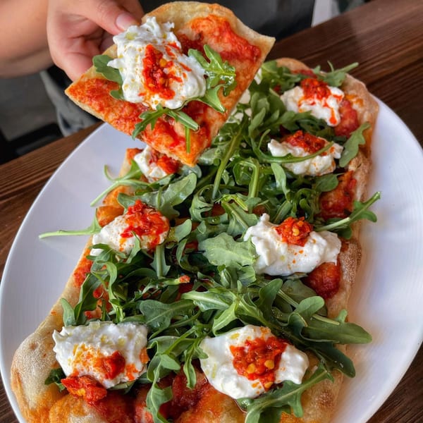 A hand holds a slice of La Bella Pinseria Romana's pinsa with burrata, arugula, and sun-dried tomatoes.
