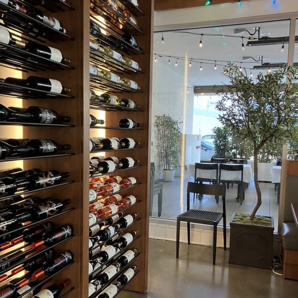 Wine racks filled with bottles next to a dining area with tables, chairs, and an olive tree.